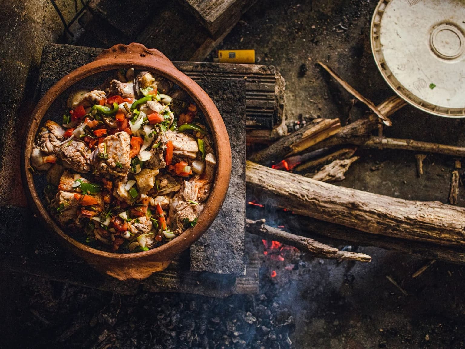 Panela de barro com carne e legumes cozinhando sobre brasas de lenha