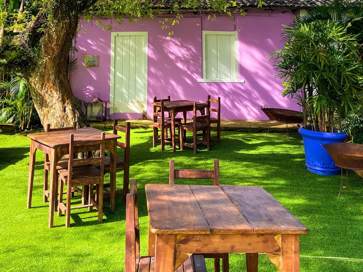 Mesas de madeira na grama, à sombra de uma árvore, diante de um casario colonial rosa no Quadrado de Trancoso